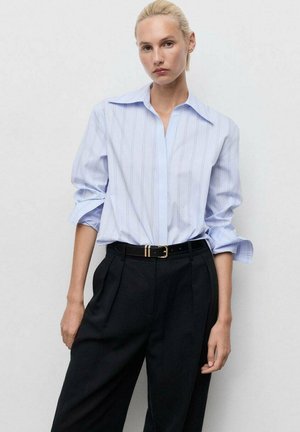 Chemise bleu clair à rayures avec un large col et des manches retroussées, associée à un pantalon noir taille haute et une fine ceinture noire.