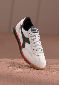 Hvit atletisk sko med mesh- og semsket skinn-materialer, svarte og røde detaljer, rund tå, snørebånd-design og teksturert gummisåle.