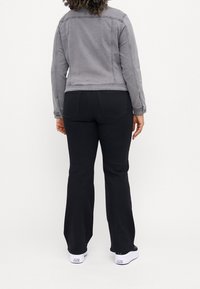 Osoba s kudrnatými vlasy, oblečená v šedé bundě, černých rozšířených kalhotách a bílých teniskách Converse, stojící zády k bílému pozadí.