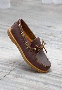 Καφέ δερμάτινο παπούτσι τύπου βάρκας με ταμπά σόλα και δερμάτινα κορδόνια δεμένα σε φιόγκο, ακουμπισμένο σε ανοιχτό γκρι πλακάκι δαπέδου.