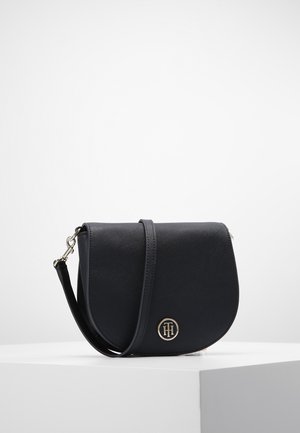 Sac à bandoulière en cuir noir avec rabat courbé et emblème circulaire doré sur le devant, posé sur un piédestal blanc sur fond uni.