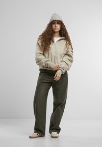 Beige zip-up hoodie met ribgebreide manchetten, gecombineerd met loszittende olijfgroene broek. Het model draagt een lichtgrijze gebreide beanie en witte sneakers.