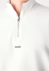 Pullover bianco con zip a un quarto e texture liscia; presenta una zip argentata e il logo "HUGO" sul petto in nero. Design minimalista.