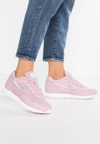 Vaaleanpunaiset urheilukengät, joissa on verkkokangas ja synteettiset materiaalit, näyttävät tekstuuri- ja muotoiluilta, valkoinen pohja ja hopeiset yksityiskohdat sivuilla.