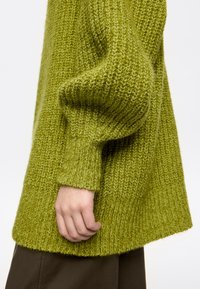 Zielony sweter wykonany na drutach o fakturowanym wykończeniu, z prążkowanymi mankietami i luźnym krojem, pokazany obok ręki spoczywającej przy dolnym brzegu.