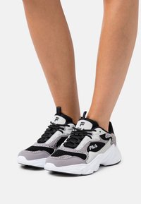 Person klädd i svart, vitt, grått och mönstrade FILA-sneakers med klumpiga vita sulor, stående mot en enkel ljus bakgrund.