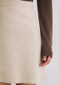 Frau trägt einen beigen Wollrock und ein braunes langärmliges Oberteil mit Daumenlöchern, steht vor einem schlichten weißen Hintergrund.