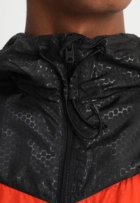 Veste noire et rouge présentant un motif hexagonal texturé avec un col haut, un cordon de serrage et une fermeture éclair. Sections en tissu lisse.