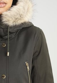 Gros plan d'un manteau d'hiver vert foncé avec une capuche bordée de fausse fourrure beige, une poche zippée dorée et des boutons-pression dorés sur le devant.