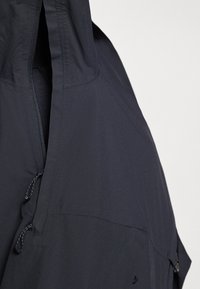Veste à capuche bleu foncé en tissu lisse et léger, avec cordons de serrage ajustables et une ouverture zippée d'un côté.