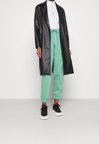 Zwarte neplederen lange jas over een witte mock neck top, gecombineerd met mintgroene joggers en zwarte sneakers met witte zolen.