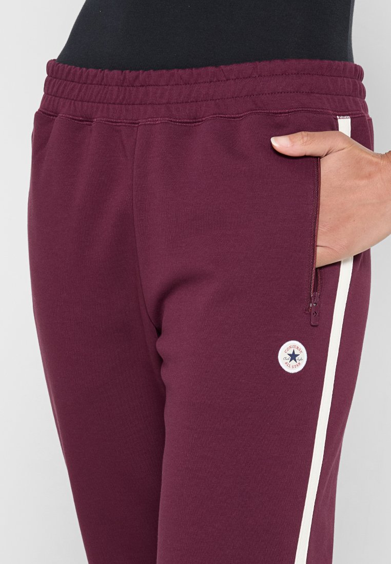 Pantalones de chándal burdeos hechos de tela suave, con una cinturilla con cordón, un bolsillo lateral con cremallera y rayas blancas en los laterales con un logo.