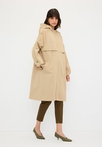 Beige regnjacka med huva, som har knappstängning, två framfickor och en avslappnad passform. Bärs över olivgröna tajta byxor.