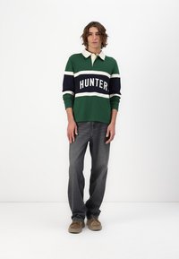 Grønn rugby skjorte med hvite og marineblå striper, med "HUNTER" tydelig trykket. Borte med grå jeans og beige fottøy.