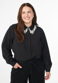 Donna sorridente con lunghi capelli castani, che indossa una blusa nera con colletto di perline bianche e pantaloni neri con coulisse, con le mani in tasca, su sfondo bianco.