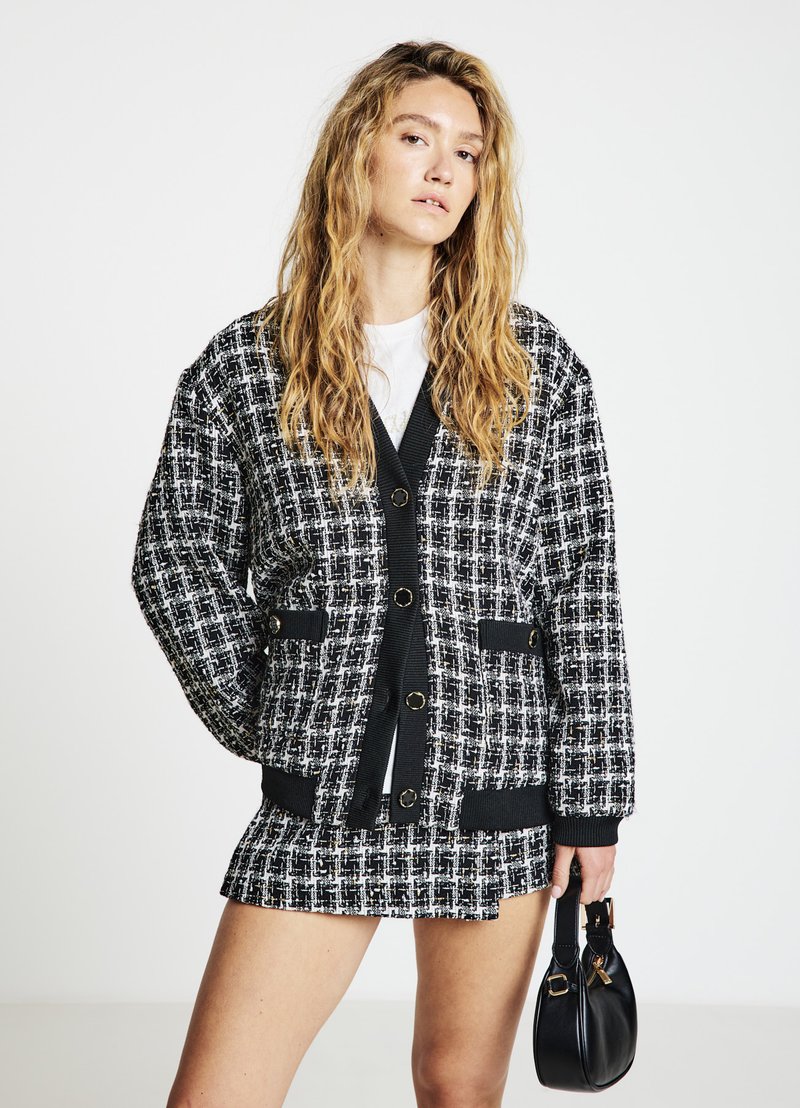 Veste en tweed noir et blanc à coupe oversize, avec deux poches avant et de gros boutons, associée à une mini-jupe assortie et un sac à main noir.