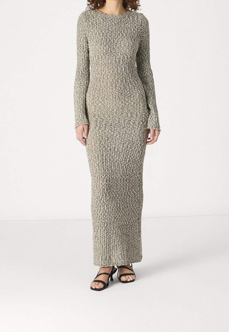 Femme portant une longue robe beige en maille côtelée à manches longues et des talons ouverts à brides noirs, debout devant un fond blanc uni.