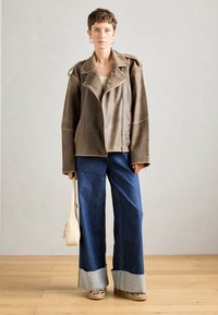 Person med kort hår iført en oversized brun skinnjakke, brede cuffed blå jeans, leopardmønstrede sko og holder en liten beige håndveske.