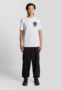 Mand står fremadskuende iført en hvid grafisk T-shirt, sorte cargobukser og sorte sneakers mod en ensfarvet grå baggrund.