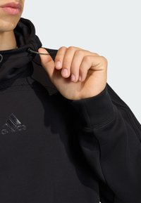 Čierna mikina s kapucňou a šnúrkou, s vyšívaným logom Adidas na hrudi. Tkanina je mäkká s hladkou textúrou.