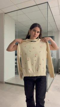 Femme aux cheveux foncés tient un pull crème avec une petite broderie florale rose dans une pièce moderne aux murs en verre.