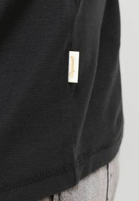 Section de tissu noir d'un vêtement avec une petite étiquette blanche portant la mention « Originals » en couture beige, attachée près de l'ourlet au-dessus d'un jean en denim gris clair.