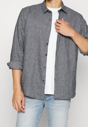 Mężczyzna noszący rozpiętą szarą koszulę na białym t-shircie i jasnoniebieskich dżinsach, z rękawami podwiniętymi do przedramion.