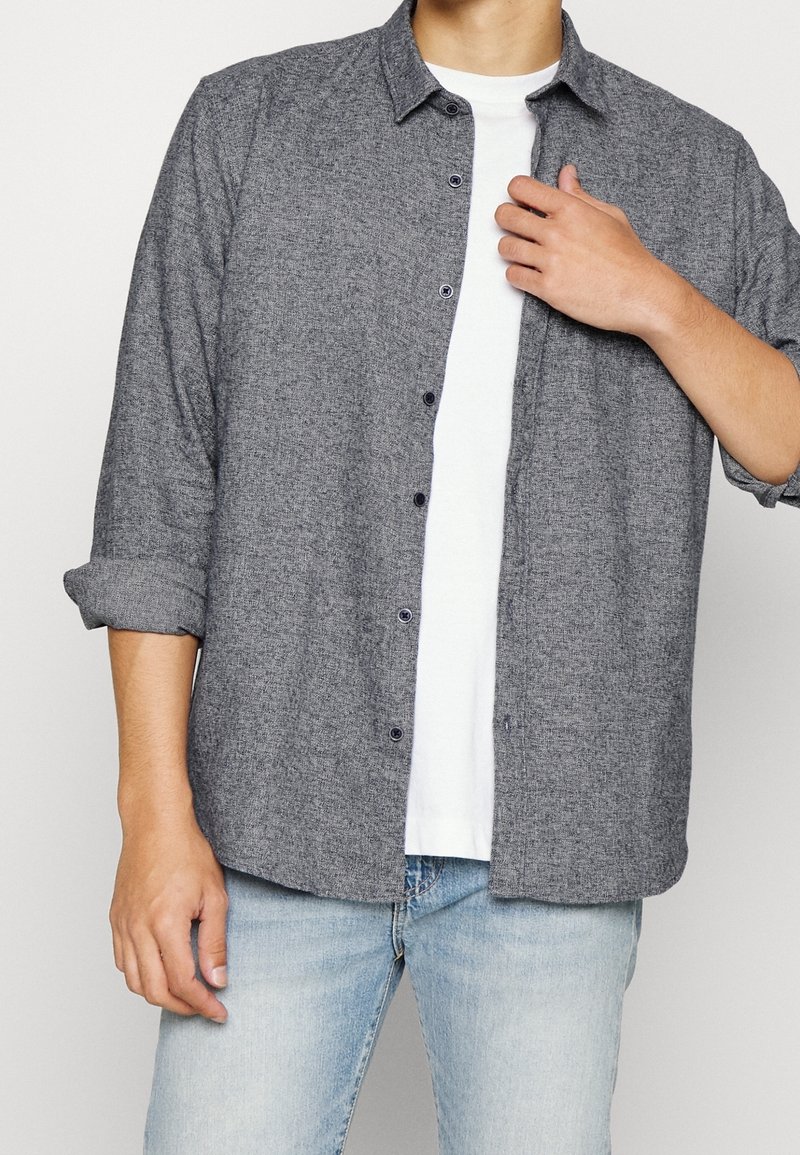 Homme portant une chemise grise déboutonnée sur un t-shirt blanc et un jean bleu clair, avec les manches retroussées jusqu'aux avant-bras.