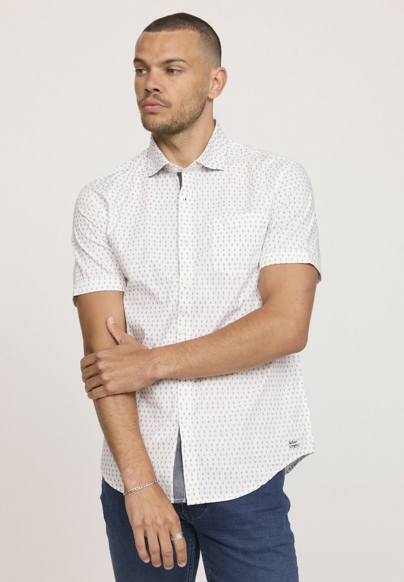 Chemise blanche à manches courtes avec un motif bleu subtil, col boutonné et poche, associée à un jean en denim foncé.
