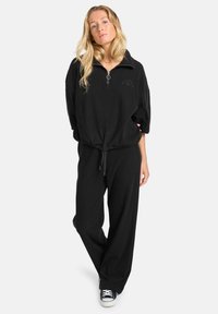 Zwarte zip-up sweatshirt met hoge kraag, gestructureerde stof en een trekkoord in de taille, gecombineerd met bijpassende wijde broek en sneakers.