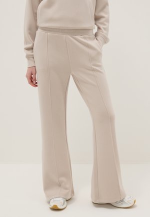 Vrouw draagt beige wijde broek en bijpassend sweatshirt met witte sneakers, staand tegen een effen lichte achtergrond.