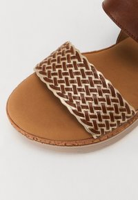 Brown leather sandal featuring a woven gold accent strap, rounded shape, and brown textured insole with a slight heel.