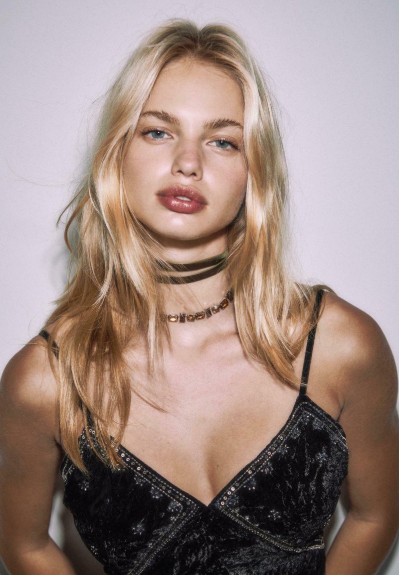 Schwarzes Samtkleid mit Perlendetails, übereinander getragene Halsketten, darunter ein dunkler Choker, und glattes, blondes Haar vor einem hellen Hintergrund.