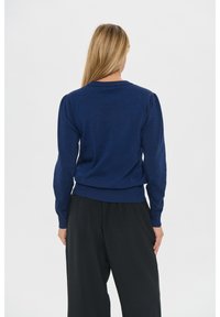 Frau mit langen blonden Haaren, die einen marineblauen Pullover mit langen Ärmeln und locker sitzende schwarze Hosen trägt, mit dem Rücken zu uns vor einem schlichten hellen Hintergrund.
