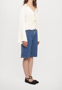 Cremefarvet, let bluse med reverskrave og bindebånd foran. Kombineret med knælange blå denimshorts. Sorte, spidse ballerinasko med prikker.