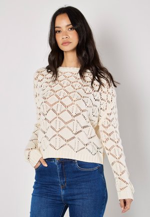 Cremfarbener, gestrickter Pullover mit einem Rautenmuster und offenen Maschen. Lange Ärmel, gerippte Bündchen und Saum. Getragen mit blauen hochtaillierten Jeans.