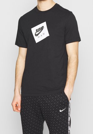 Camiseta de algodón negra con un logo en un recuadro blanco que dice "JUMP" y "MAN", combinada con pantalones de chándal negros con un patrón y un logo lateral.