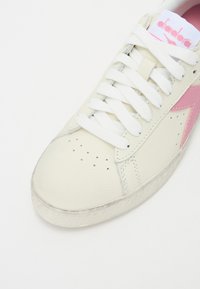 Baskets en cuir blanc avec une accent rose, lacets blancs, boîte à orteils perforée et semelle en caoutchouc texturée. Logo Diadora sur la languette.