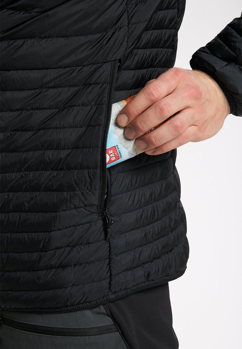 Chaqueta aislante negra con textura acolchada, que presenta un bolsillo con cremallera donde se inserta parcialmente una barra de energía colorida.
