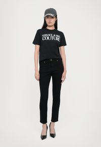 Juodas džinsinis marškinėlis su baltu "VERSACE JEANS COUTURE" spaudiniu, derinamas su juodais prigludusiais džinsais ir juodais siaurais batuotais kulniukais. Pilka kepurė akcentui.