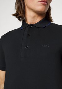 Černé polo tričko vyrobené z texturované tkaniny, s dvouknoflíkovou légou, jemnými modrými akcenty na límci a vyšitým logem „BOSS“ na hrudi.