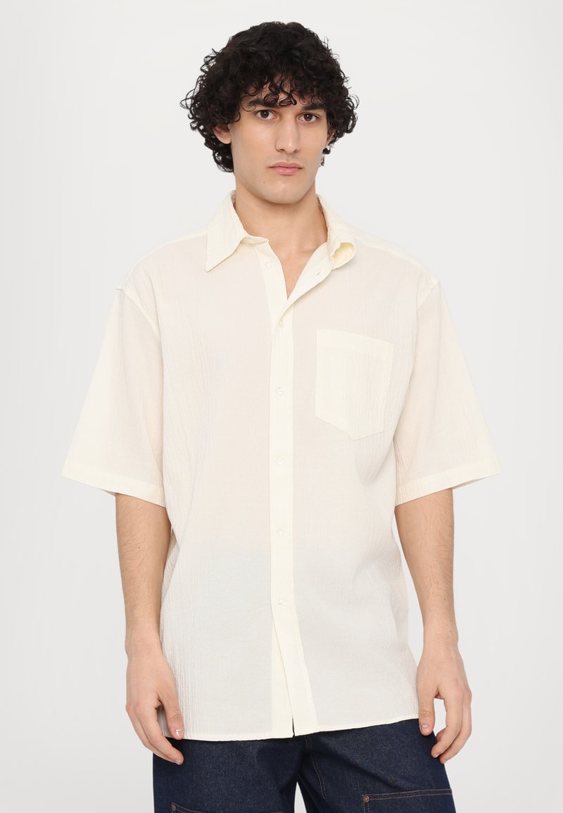 Camisa de botón de manga corta en tejido texturizado color blanco roto, con un bolsillo en el pecho y cuello clásico, que presenta un corte relajado y dobladillo recto.