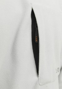 Fleece jakna u boji off-white s mekanom teksturom, s džepom na zip i detaljima od crnog zamka, s suptilnim brendingom na tkanini.