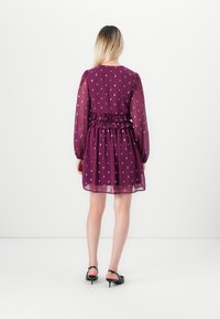 Kvinna klädd i en plommonfärgad klänning med silverprickar, långa genomskinliga ärmar, rynkad midja och svarta kitten heels slingback skor, stående mot en vit vägg.