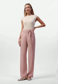 Pantalones de pierna recta de color rosa claro con un detalle de cintura anudada, combinados con una camiseta de canalé ceñida en color crema. Modelo con sandalias planas metálicas.