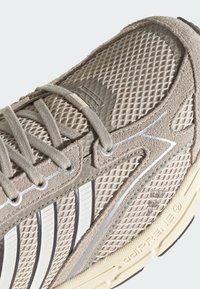 Nahaufnahme eines beigen Mesh- und Veloursledersneakers mit grauen Schnürsenkeln, weißen Streifen und strukturierter Sohle mit der Aufschrift "adiPRENE+".