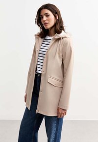 Beige vattentålig jacka med huva, framzipper och tryckknappsfickor. Bärs över en randig skjorta och ihop med vida jeans.
