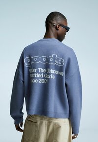 Man in een blauwe trui met de tekst "goods Wear The Unknown Untitled Goods since 2001" op de achterkant, beige broek en zwarte zonnebril, met zijn rug naar de kijker gekeerd.