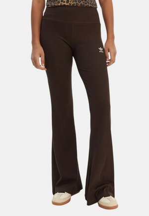 Persona che indossa pantaloni svasati Adidas marrone scuro con logo sulla coscia e sneakers beige, in piedi davanti a uno sfondo bianco.