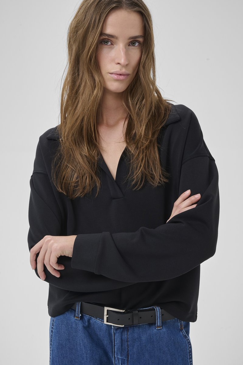 Donna con capelli castano chiaro lunghi, indossa una maglia nera a maniche lunghe e jeans blu, in piedi con le braccia incrociate davanti a uno sfondo chiaro e uniforme.